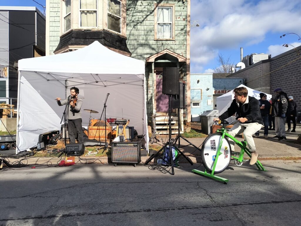 Pedal powered stage. A person is playing violin and another person is riding a stationary bike that is powering a speaker. 